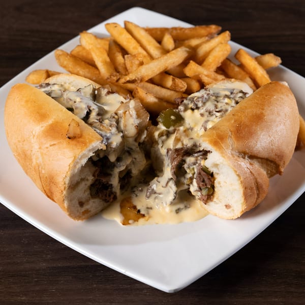 A cheesesteak sandwich, cut in half and overflowing with melted cheese, onions, and peppers, served with fries.