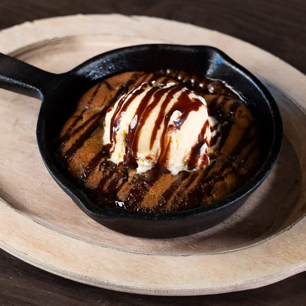 Warm chocolate chip cookie skillet with vanilla ice cream and chocolate drizzle on a wooden tray.