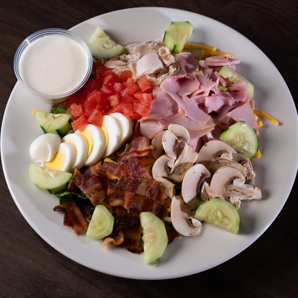 A large chef salad with ham, turkey, bacon, tomatoes, mushrooms, hard-boiled eggs, and ranch dressing.