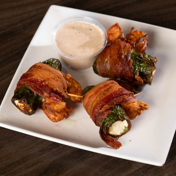 Bacon-wrapped jalapeño poppers with a side of dipping sauce on a white rectangular plate.